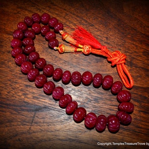 May include: A red gemstone mala with a red and gold tassel. The mala is made of 108 beads and is strung on a red cord.