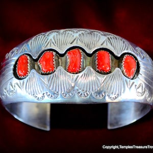 May include: A wide, silver cuff bracelet with five vibrant red coral stones set in individual bezels. The bracelet features intricate, stamped patterns along the top edge, with a smooth, polished finish. The open design allows for easy wear.