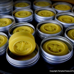 May include: A collection of round, yellow, honey-themed soaps in metal tins. The soaps have a bee design and are arranged in a variety of sizes and positions.