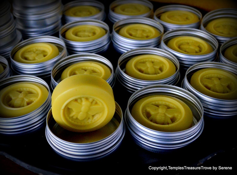 May include: A collection of round, yellow, honey-themed soaps in metal tins. The soaps have a bee design and are arranged in a variety of sizes and positions.