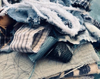 May include: A close-up of a collection of fabric scraps in various shades of blue, white, and tan. The fabrics include denim, patterned cotton, and textured weaves. White, frayed yarn is draped over the top, adding a tactile element. The arrangement suggests a craft project or textile art.