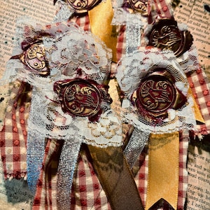 May include: Several handmade brooches with a vintage aesthetic. Each brooch features a round, embossed metal button with a bird design, layered with lace, ribbons, and a red and white checkered fabric.