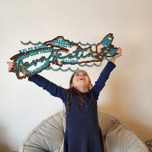May include: A young girl holds up a wooden cutout of the province of Nova Scotia, with the names of various cities and towns written on it. The cutout is painted in shades of brown and blue.