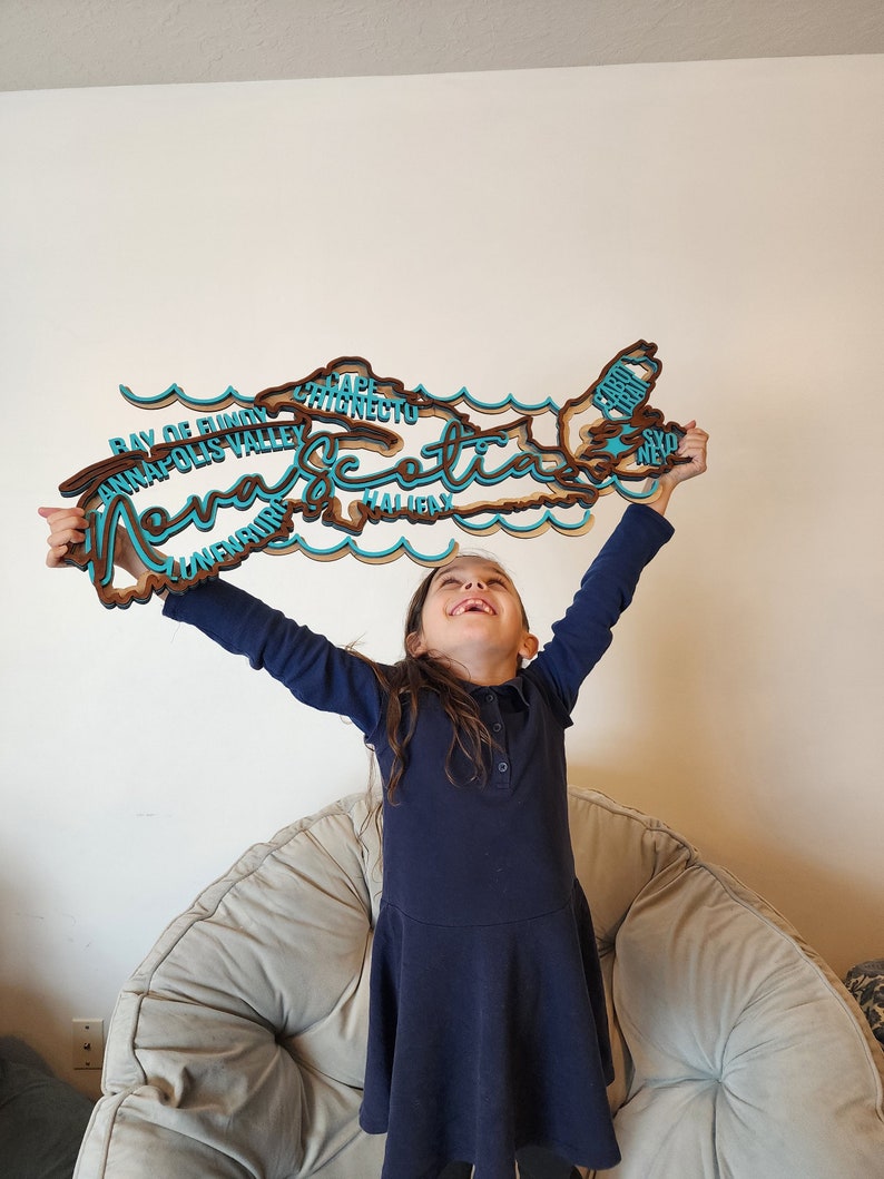 May include: A young girl holds up a wooden cutout of the province of Nova Scotia, with the names of various cities and towns written on it. The cutout is painted in shades of brown and blue.