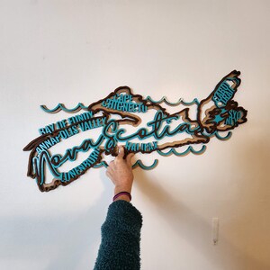 May include: A wooden cutout of the province of Nova Scotia, painted in brown and teal blue. The outline of the province is filled with the names of cities and towns, including Halifax, Cape Breton, and Sydney.