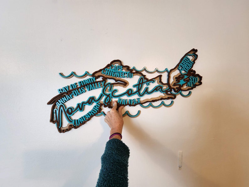 May include: A wooden cutout of the province of Nova Scotia, painted in brown and teal blue. The outline of the province is filled with the names of cities and towns, including Halifax, Cape Breton, and Sydney.