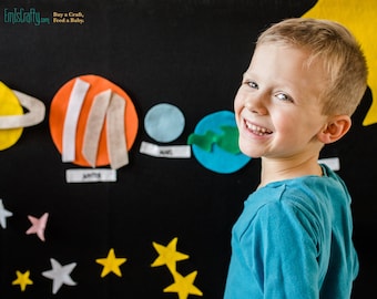 May include: A young boy smiles while standing in front of a felt solar system wall art. The art features planets, stars, and the text "Buy a Craft, Feed a Baby."