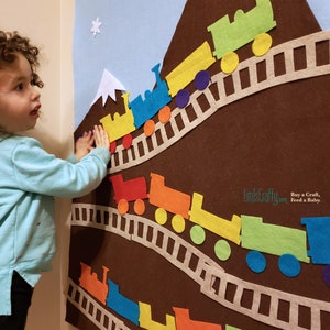 May include: A felt play set with a brown mountain and a train track with colorful trains going up the mountain. The train cars are yellow, red, orange, green, blue, and purple. The track is made of felt and has a white border. The background is a light blue sky with a white cloud and snowflake.