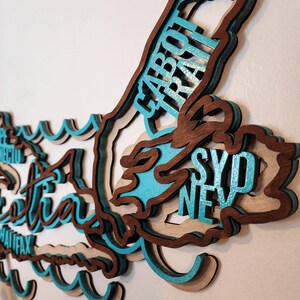 May include: A wooden wall art piece shaped like the island of Nova Scotia, Canada. The island is layered and painted in shades of brown and teal. The names of cities on the island are written in white lettering, including Sydney, Halifax, and Cape Breton.
