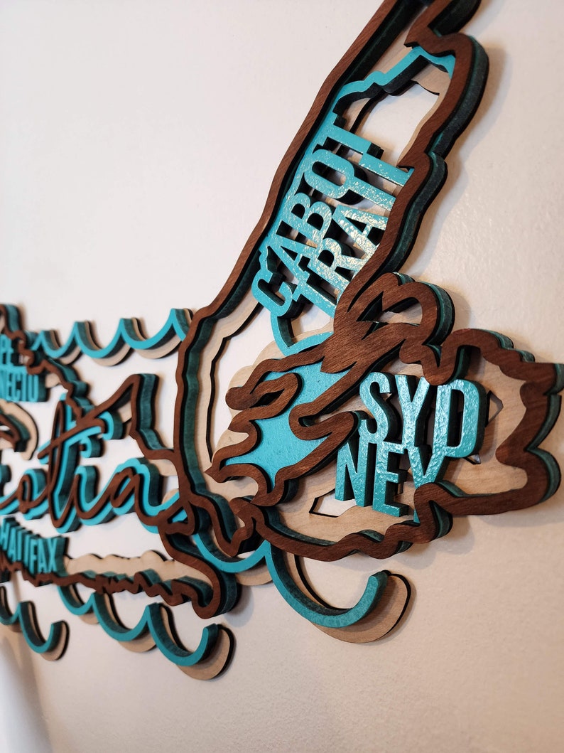 May include: A wooden wall art piece shaped like the island of Nova Scotia, Canada. The island is layered and painted in shades of brown and teal. The names of cities on the island are written in white lettering, including Sydney, Halifax, and Cape Breton.