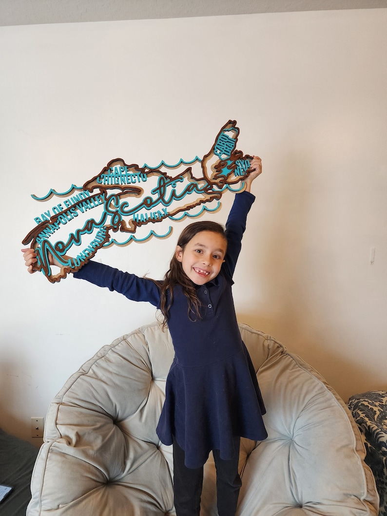 May include: A young girl wearing a navy blue dress holds up a wooden cutout of the province of Nova Scotia. The cutout is painted in brown and teal colours and features the names of various cities and regions within the province.