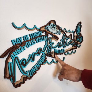 May include: A wooden wall art piece shaped like the province of Nova Scotia, Canada. The outline of the province is filled with the names of cities and towns in Nova Scotia, including Halifax, Lunenburg, Annapolis Valley, and Cape Chignecto. The piece is painted in shades of brown and blue.