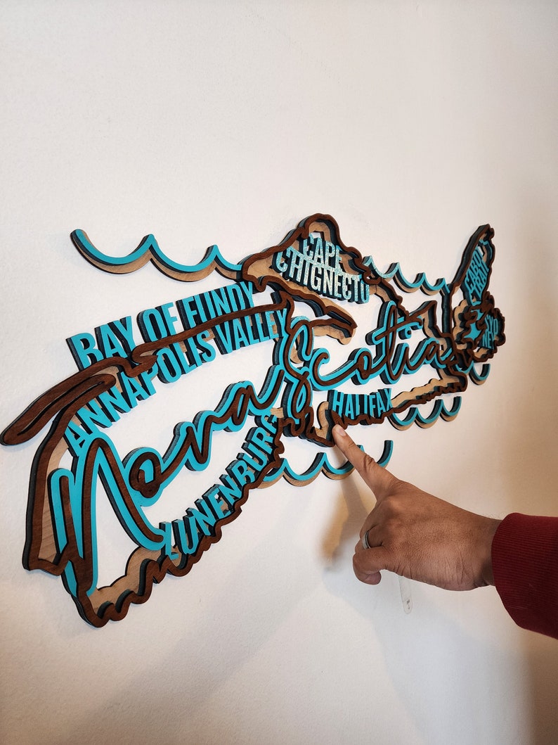 May include: A wooden wall art piece shaped like the province of Nova Scotia, Canada. The outline of the province is filled with the names of cities and towns in Nova Scotia, including Halifax, Lunenburg, Annapolis Valley, and Cape Chignecto. The piece is painted in shades of brown and blue.
