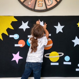 Puede incluir: Un niño coloca una estrella blanca de fieltro en una pizarra de fieltro negro con un sol amarillo, planetas y otras estrellas. La pizarra está decorada con un tema espacial.