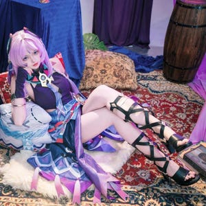 Op de afbeelding: Een persoon in een paars en blauw cosplaykostuum met een pruik, handschoenen en sandalen. Het kostuum heeft ingewikkelde details en is versierd met sieraden. De persoon ligt op een kleed met patronen met kussens.