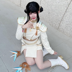 Może przedstawiać: Biały kostium cosplay z złotymi akcentami, w tym naramienniki, krótki top i spódnica. Strój jest połączony z białymi butami i czarną peruką w stylu koków. Widoczne są również ozdobne akcesoria do włosów oraz niebieskie i złote ornamenty.