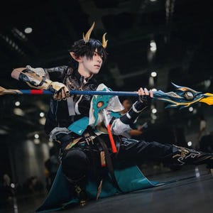 Könnte beinhalten: Eine Person in einem detaillierten Cosplay-Kostüm mit schwarzen und türkisfarbenen Akzenten hält einen Stab. Das Kostüm umfasst goldene Hörner, komplizierte Muster und einen fließenden türkisfarbenen Umhang. Der Stab hat ein blaues und goldenes Design.