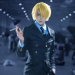 Könnte beinhalten: Eine Person in einem Cosplay-Outfit, die einen dunkelblauen, zweireihigen Anzug mit goldenen Knöpfen, ein hellblau gestreiftes Hemd und eine schwarze Krawatte trägt. Die Person hat kurze, stachelige blonde Haare und hält eine brennende Zigarette.