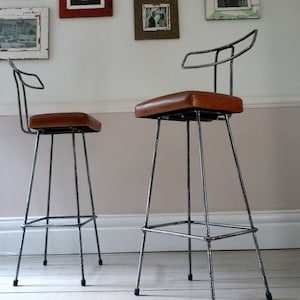May include: Two industrial-style bar stools with a metal frame and brown leather seats. The stools have a simple design with a curved backrest and a footrest.