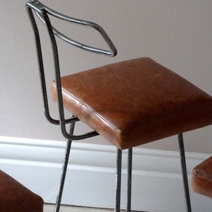 May include: Two metal bar stools with brown leather seats. The stools have a simple, modern design with a curved backrest.