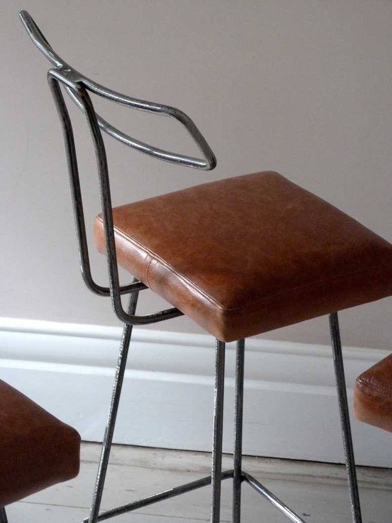 May include: Two metal bar stools with brown leather seats. The stools have a simple, modern design with a curved backrest.