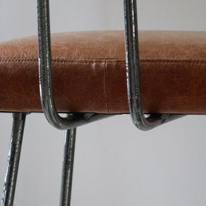 May include: Close-up of a brown leather chair seat with a metal frame. The chair has a simple, modern design with thin metal legs.
