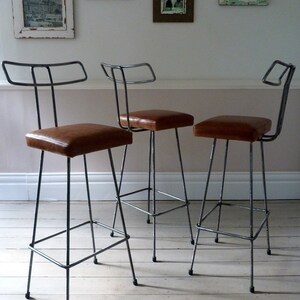 May include: Three metal bar stools with brown leather seats. The stools have a simple, industrial design and are perfect for a modern kitchen or dining room.