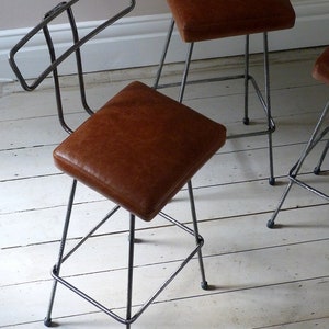 May include: Two brown leather bar stools with a metal frame. The stools have a simple, industrial design with a backrest and four legs.