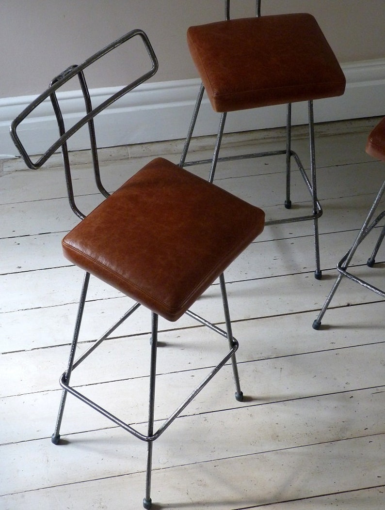 May include: Two brown leather bar stools with a metal frame. The stools have a simple, industrial design with a backrest and four legs.
