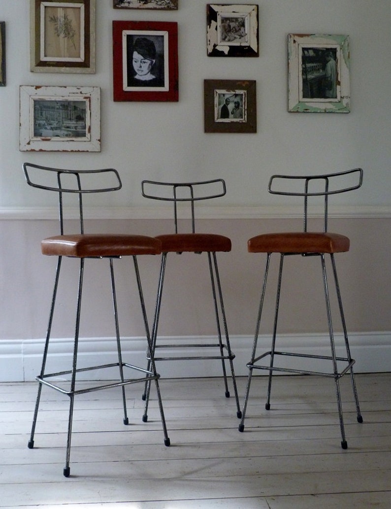 May include: Three black metal bar stools with brown leather seats. The stools have a simple, industrial design and are arranged in a row on a white wooden floor.