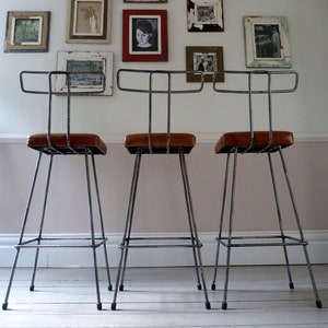 May include: Three black metal bar stools with brown leather seats. The stools have a simple, industrial design and are arranged in a row.