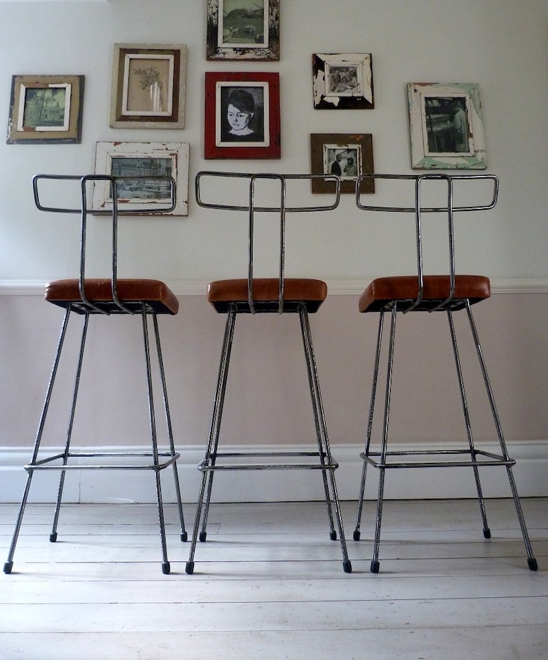 May include: Three black metal bar stools with brown leather seats. The stools have a simple, industrial design and are arranged in a row.