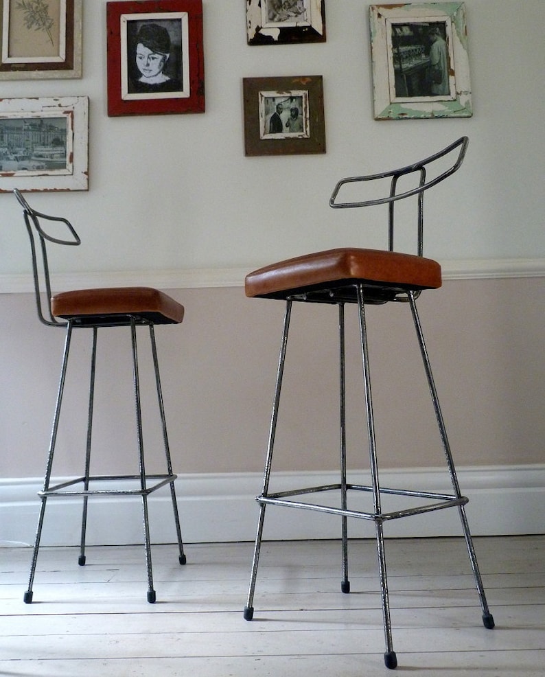 May include: Two industrial-style bar stools with a metal frame and brown leather seats. The stools have a simple design with a curved backrest and a footrest.