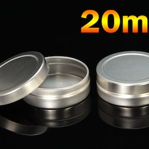 Può includere: Tre piccoli barattoli rotondi in metallo argentato con coperchi. Un coperchio è rimosso, rivelando l'interno del barattolo. Il testo "20ml" è visualizzato in arancione e giallo sopra i barattoli. I barattoli sono su una superficie nera riflettente.