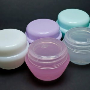 以下が含まれることがあります： 蓋付きの小さな丸いプラスチック容器が5つあります。容器は白、薄い紫、薄い青、ピンクがかった透明など、さまざまな色です。