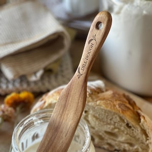 Peut inclure: Un racloir à pâte en bois avec les mots "Dill Dough" gravés sur le manche. Le racloir est posé dans un bocal en verre contenant un levain blanc. Une miche de pain au levain se trouve à l'arrière-plan.