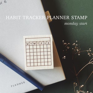 May include: A wooden stamp with a grid pattern and the letters M, T, W, T, F, S, S. The stamp is on a white planner with the word "PLANNER" printed on it. The background is a dark green surface.