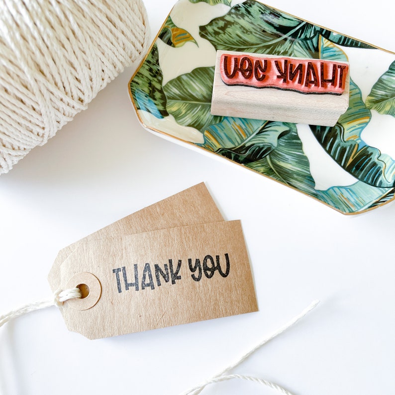 May include: A craft supplies still life featuring a brown tag with the words "THANK YOU" printed in black, a wooden stamp with the word "THANK YOU" in red, a decorative tray with a green leaf pattern, and a roll of white twine.