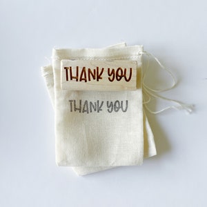 May include: A wooden stamp with the words "THANK YOU" in a handwritten font rests on a small, off-white drawstring bag. The same phrase is printed in gray on the bag. The stamp and bag are on a white surface.