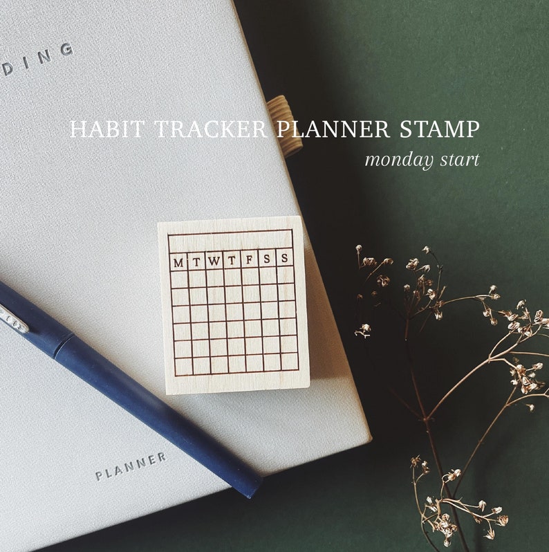 May include: A wooden stamp with a grid pattern and the letters M, T, W, T, F, S, S. The stamp is on a white planner with the word "PLANNER" printed on it. The background is a dark green surface.