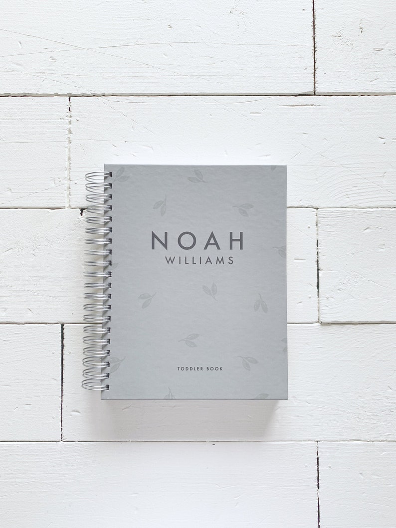 May include: A gray spiral bound book with a light gray background and a subtle leaf pattern. The book has the name "Noah Williams" and the text "Toddler Book" printed on the front cover.