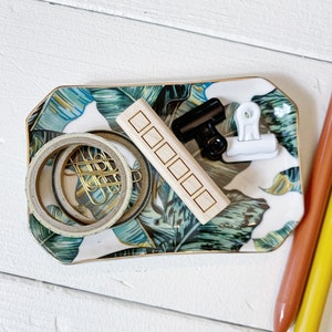 May include: A white ceramic tray with a green and brown tropical leaf pattern. The tray contains a wooden block with five squares carved into it, two gold paper clips, a black binder clip, a white binder clip, and two rolls of washi tape.