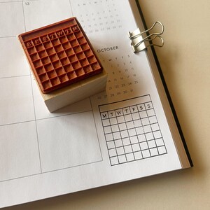 May include: A wooden stamp with a brown rubber calendar grid. The stamp is labeled "220TWTM" and is used to mark dates on a calendar.