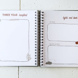 May include: A white journal with a brown spiral binding. The left page has the text "THREE YEAR handprint" and a blank space for a handprint. The right page has the text "highs and lows" and two blank spaces for writing.
