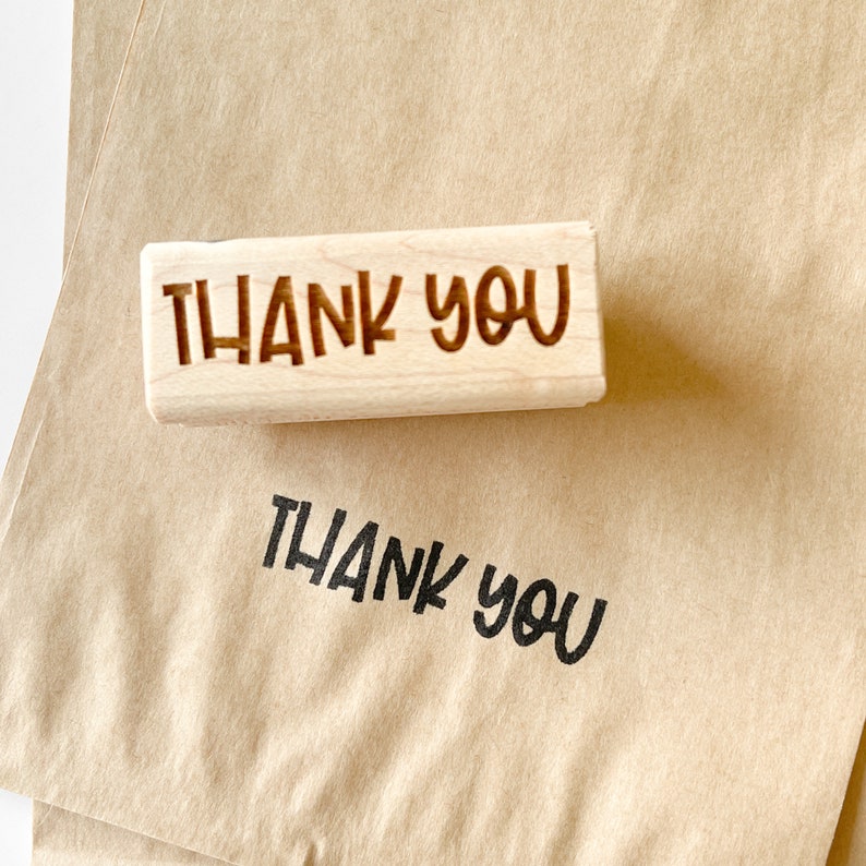 May include: A wooden stamp with the words "THANK YOU" in a handwritten font is shown on a brown paper surface. The stamped impression of "THANK YOU" is also visible below the stamp.