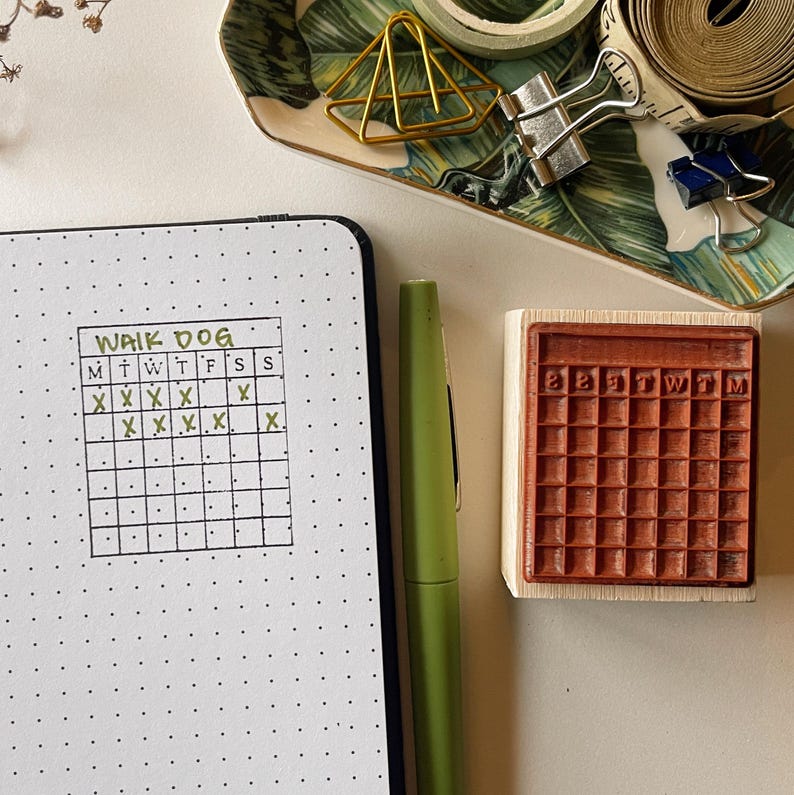 May include: A black and white planner with a green handwritten heading "Walk Dog" and a gridded section with "M T W T F S S" labeled across the top. The grid has "X" marks in some of the squares. A wooden stamp with a grid pattern is next to the planner.
