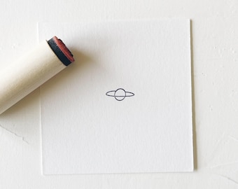 May include: A wooden stamp with a black and red handle. The stamp is used to create a simple line drawing of a planet with rings.