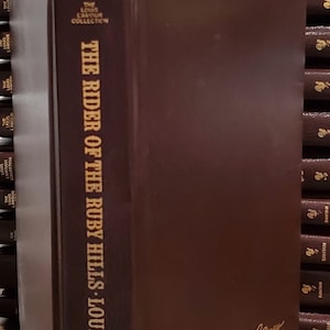 May include: A brown hardcover book with gold lettering. The title is "The Rider of the Ruby Hills" by Louis L'Amour. The book is part of "The Louis L'Amour Collection".
