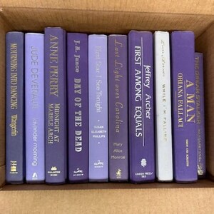 Puede incluir: Una colecci&oacute;n de diez libros de tapa dura de color morado y azul con letras doradas en los lomos. Los libros est&aacute;n dispuestos verticalmente en una fila sobre una superficie marr&oacute;n.