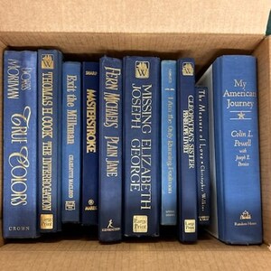 Puede incluir: Una colecci&oacute;n de 12 libros de tapa dura azules con letras doradas. Los libros est&aacute;n dispuestos verticalmente en una caja de cart&oacute;n. Los t&iacute;tulos de los libros incluyen "True Colors", "The Interrogation", "Exit the Milkman", "Masterstroke", "Plain Jane", "Missing Elizabeth", "I Am He, Only Running Freedom", "The Measure of Love" y "My American Journey".